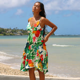 A woman wearing a vibrant tropical floral print dress stands on a sandy beach with the ocean and blue sky in the background.