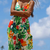 A woman wearing a sleeveless V-neck dress with a vibrant tropical floral print in red, orange, yellow, green, and blue on a cream background, photographed outdoors against a blue sky with clouds.