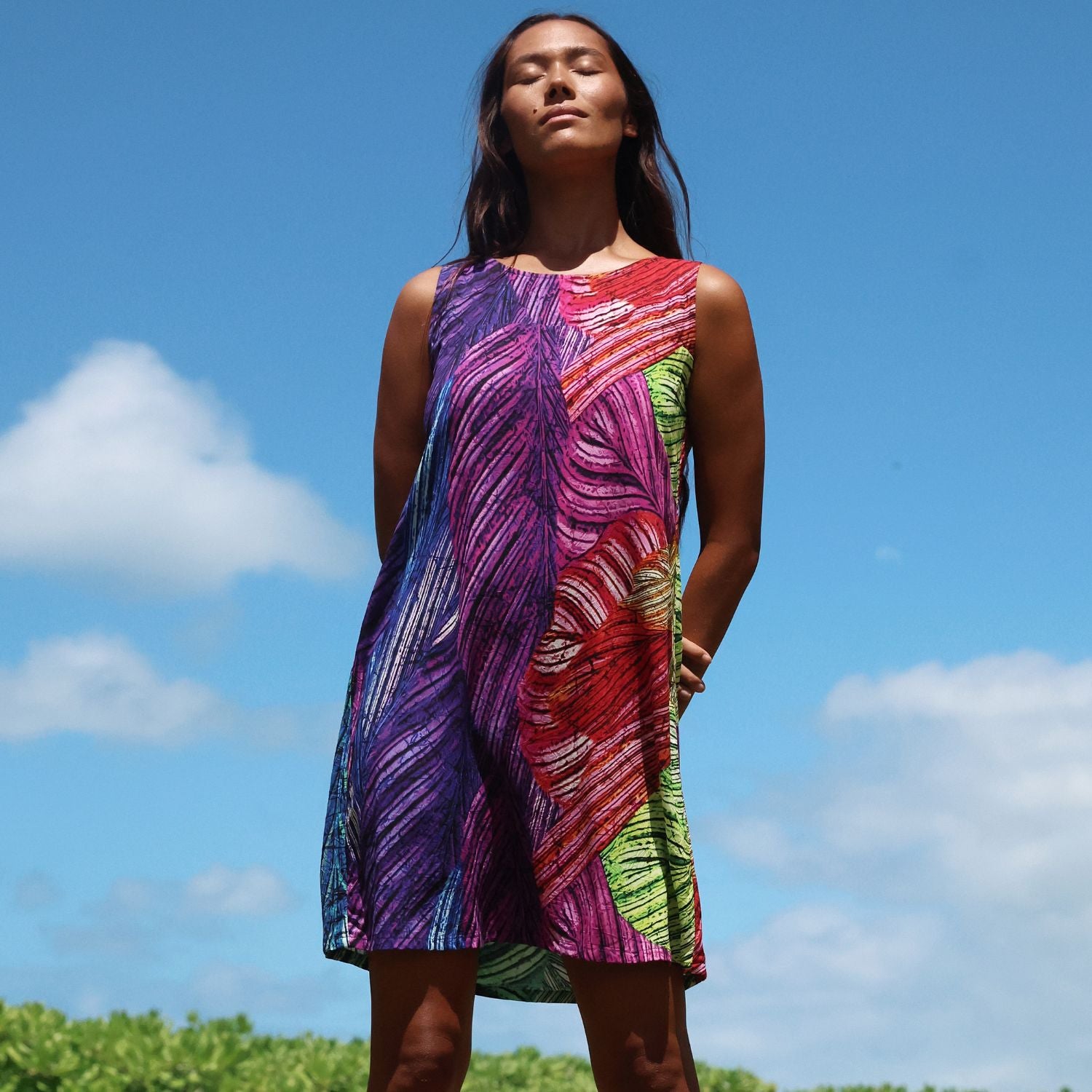 A woman wearing a sleeveless, knee-length dress with a vibrant multicolor abstract leaf-like print, standing outdoors against a bright blue sky with clouds.