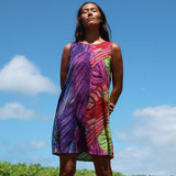 A woman wearing a sleeveless, knee-length dress with a vibrant multicolor abstract leaf-like print, standing outdoors against a bright blue sky with clouds.