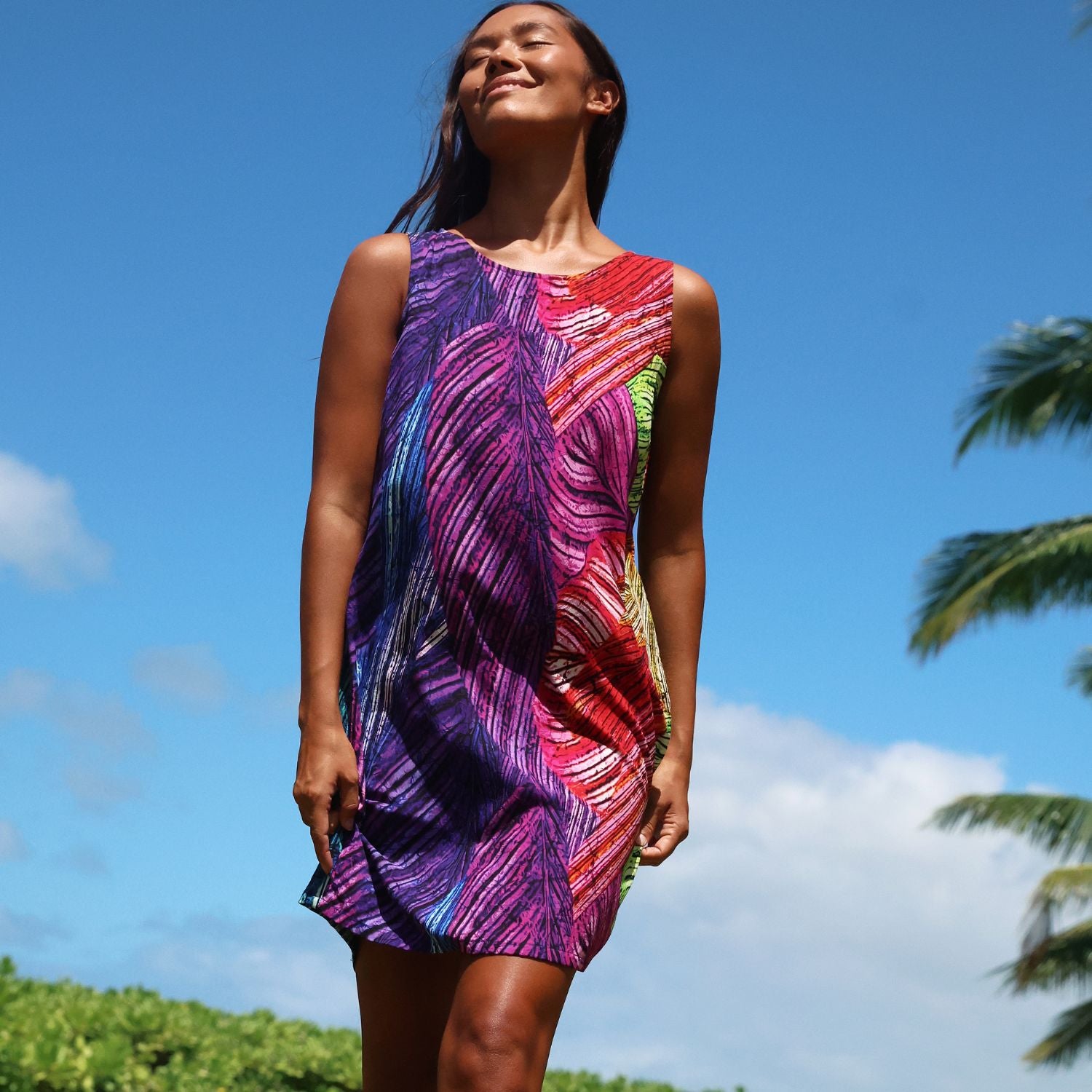 A woman wearing a sleeveless, knee-length dress with a vibrant multicolor abstract leaf pattern in purple, pink, red, and green, photographed outdoors against a bright blue sky with palm trees.