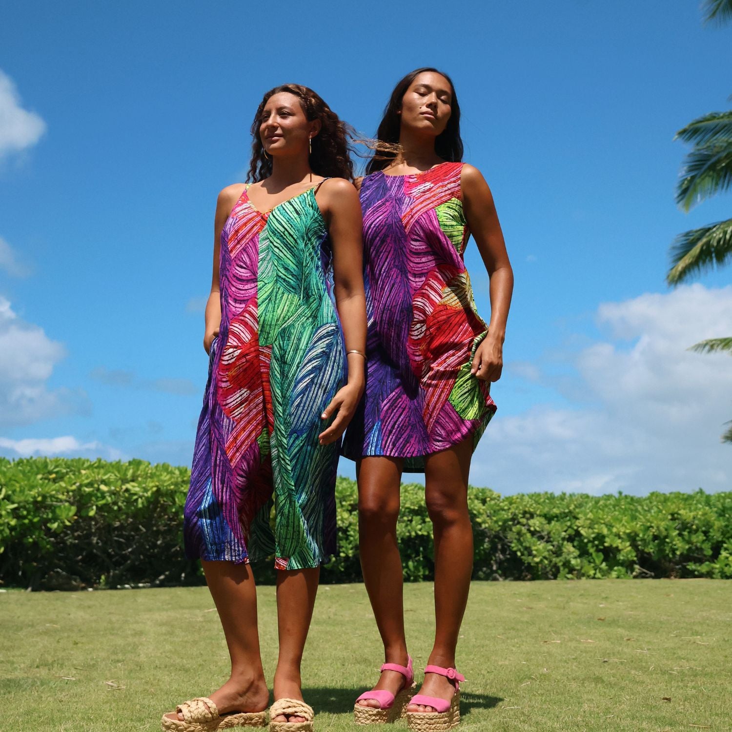 Two women standing outdoors on a grassy lawn under a blue sky, wearing matching sleeveless dresses with a bold multicolor abstract leaf-like pattern in purple, green, red, and blue.