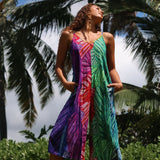 A woman wearing a vibrant, multicolor sleeveless dress with a leaf print pattern, standing outdoors against a backdrop of palm trees and sky.