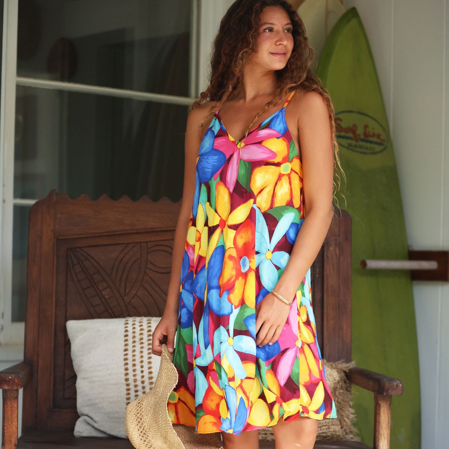 Una mujer con un vestido sin mangas de estampado floral colorido está de pie en un porche sosteniendo un sombrero de paja. Al fondo se ven una silla de madera y una tabla de surf verde.