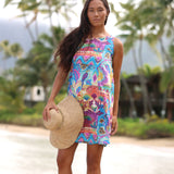 A woman wearing a colorful sleeveless tropical print dress and holding a straw hat, standing outdoors on a beach path with palm trees in the background.