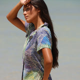 Una mujer con una camisa de manga corta abotonada con un estampado abstracto azul, verde y blanco, de pie al aire libre contra un fondo gris claro difuminado.