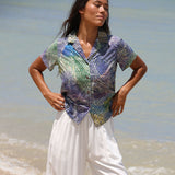 Una mujer está de pie en una playa vistiendo una camisa de manga corta abotonada con un estampado abstracto multicolor en azul, verde, morado y amarillo, combinada con pantalones blancos holgados.