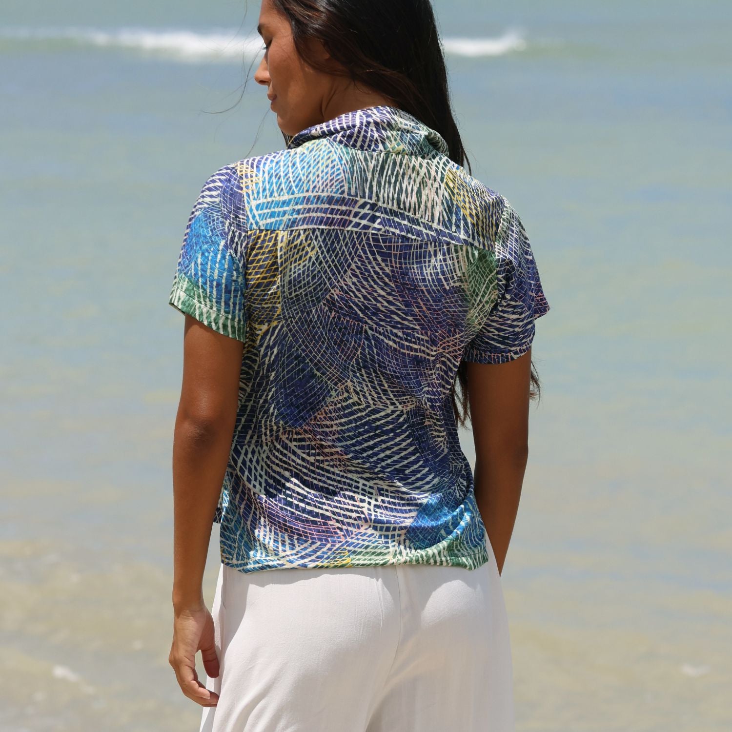 Una mujer de pie en una playa, vista desde atrás, lleva una camisa de manga corta con cuello y un colorido estampado abstracto en azul, verde, morado y blanco, combinada con pantalones blancos.