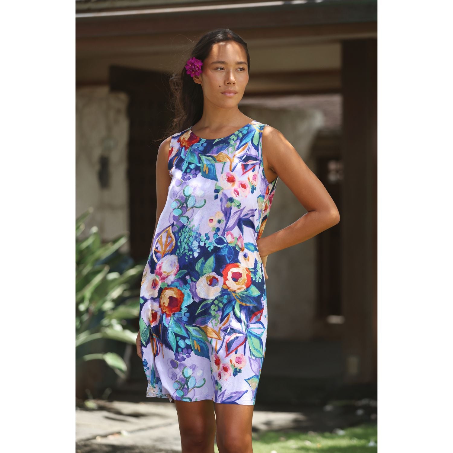 A woman wearing a sleeveless, knee-length shift dress with a vibrant multicolor floral print on a white base, standing outdoors in front of a porch.