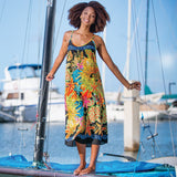A woman stands barefoot on a sailboat in a marina, wearing a colorful sleeveless maxi dress with a vibrant tropical floral print on a black background.