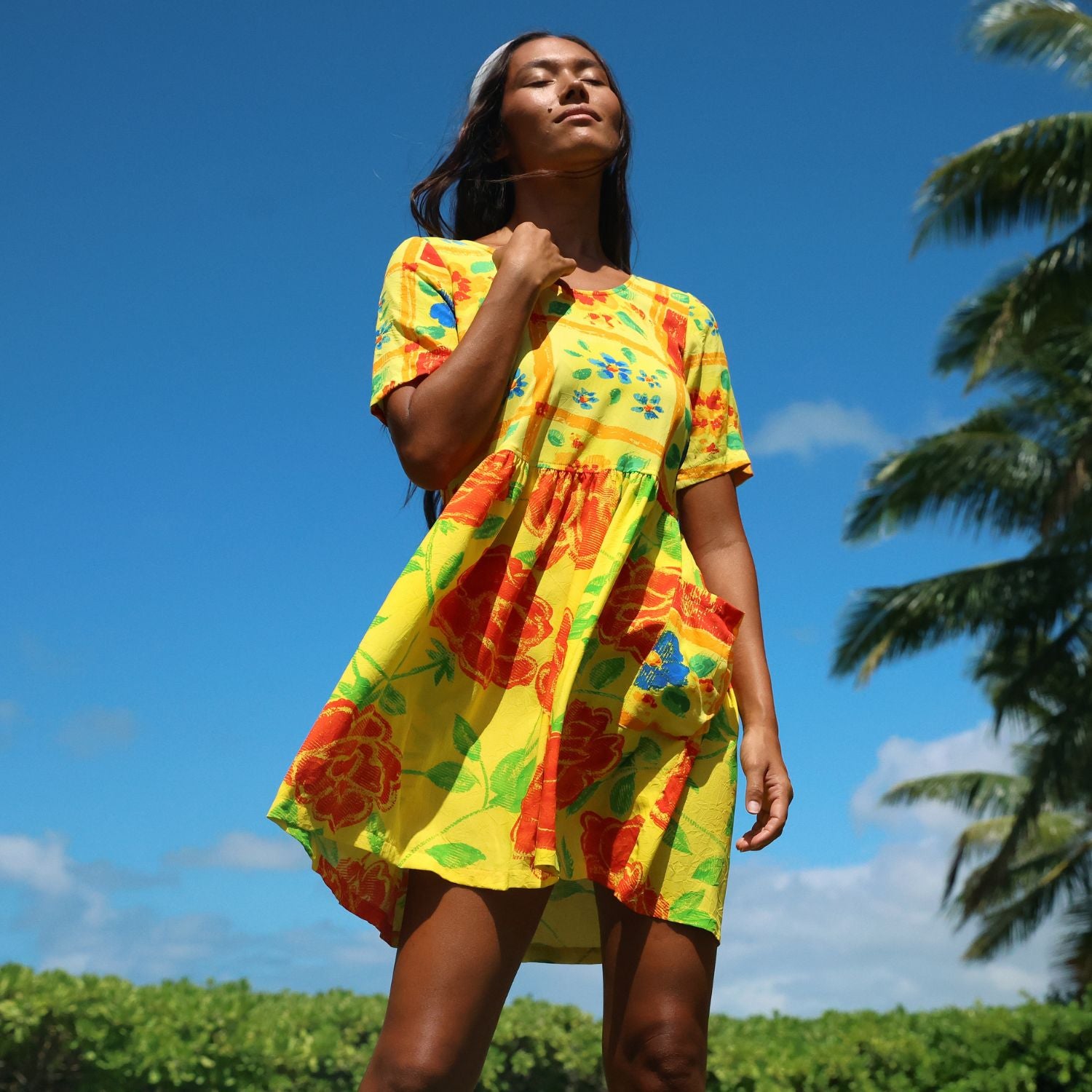 A woman wearing a bright yellow short-sleeve dress with a red, orange, and green floral print, photographed outdoors against a blue sky and palm trees.