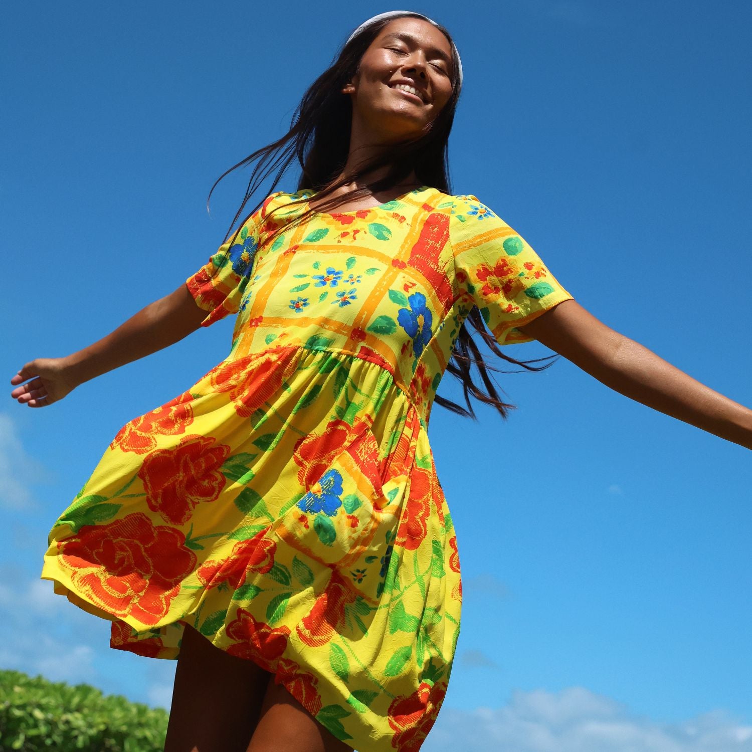 A woman wearing a bright yellow short-sleeve dress with a colorful floral and patchwork print, captured from a low angle against a clear blue sky.