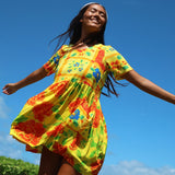 A woman wearing a bright yellow short-sleeve dress with a colorful floral and patchwork print, captured from a low angle against a clear blue sky.