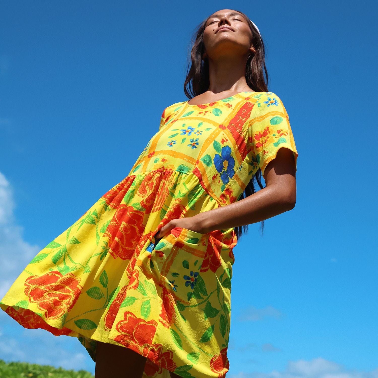 A woman wearing a bright yellow tiered dress with a red, orange, and blue floral patchwork print, photographed from a low angle against a clear blue sky.