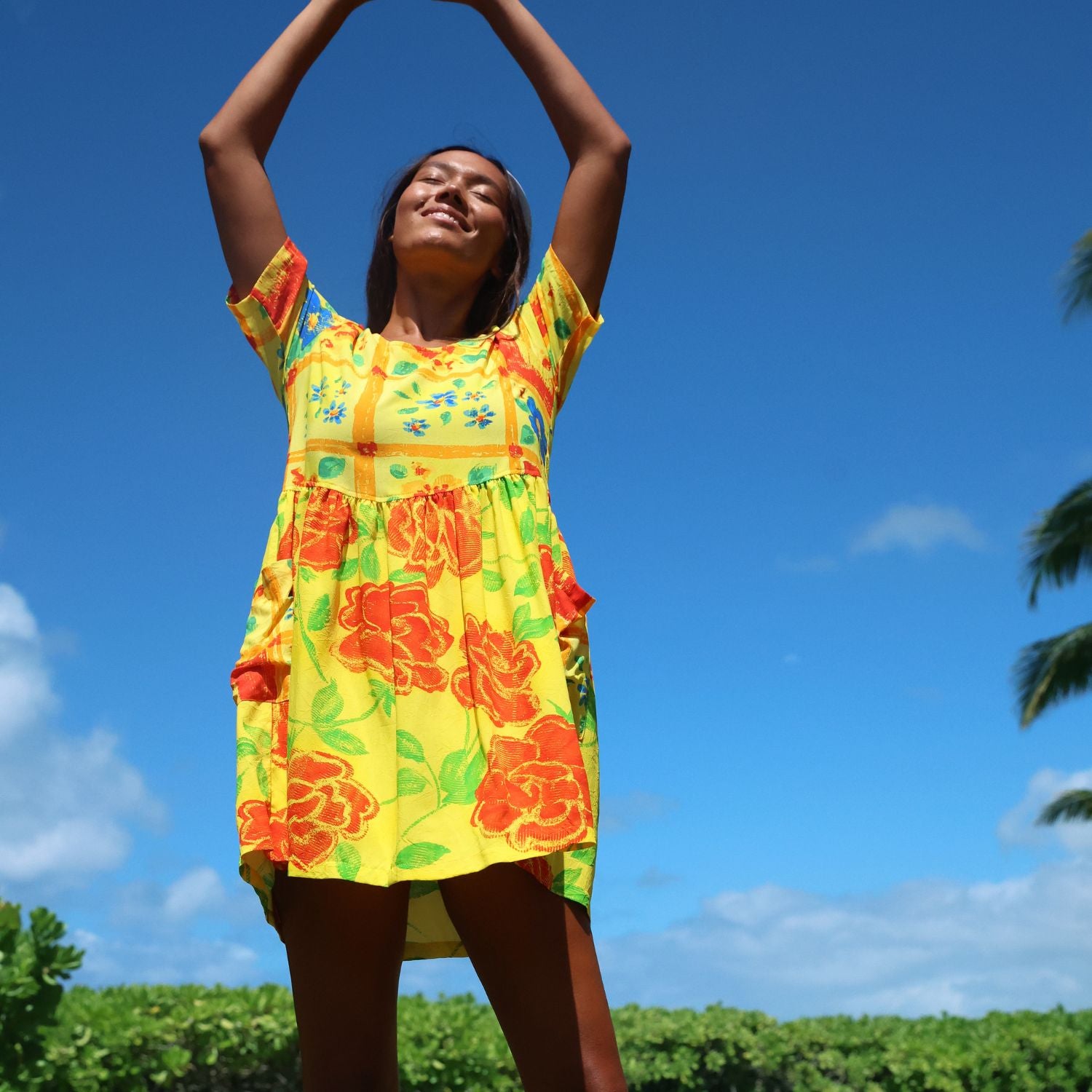A woman wearing a bright yellow short-sleeve dress with a bold orange and green floral print, posing outdoors with her arms raised against a clear blue sky.