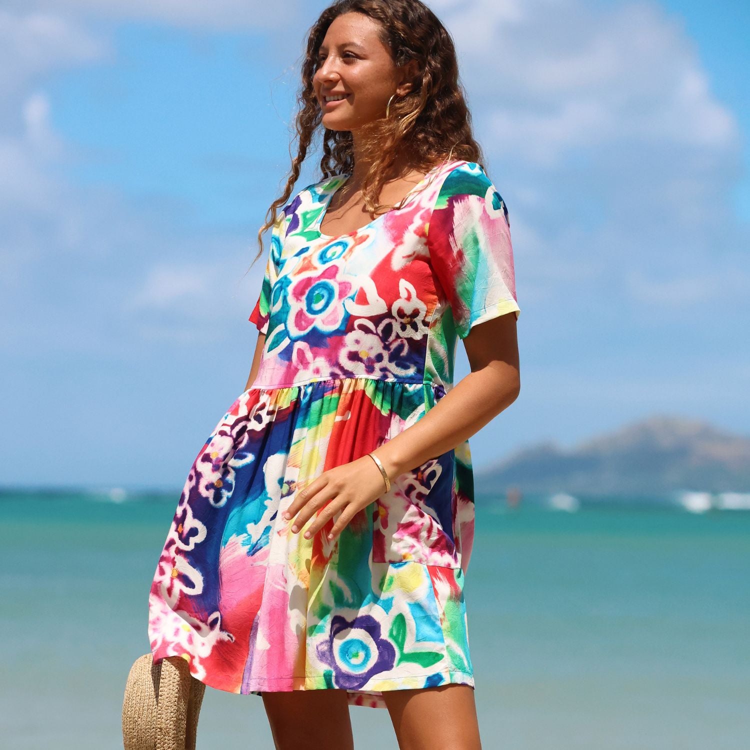 A woman wearing a colorful short-sleeve V-neck dress stands on a beach with the ocean and sky in the background. The dress features a vibrant multicolor floral and rainbow pattern.