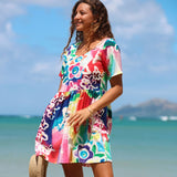 A woman wearing a colorful short-sleeve V-neck dress stands on a beach with the ocean and sky in the background. The dress features a vibrant multicolor floral and rainbow pattern.
