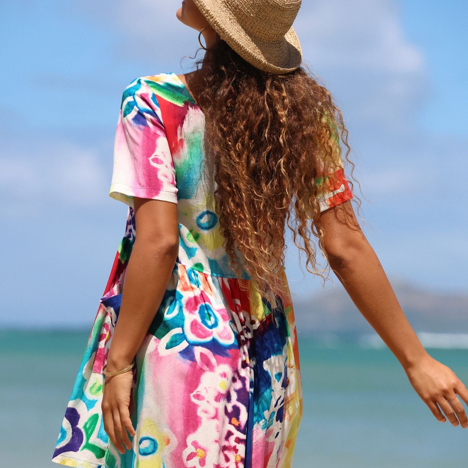 A woman wearing a colorful short-sleeve dress and a straw hat, photographed from behind on a beach with the ocean and blue sky in the background.