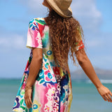A woman wearing a colorful short-sleeve dress and a straw hat, photographed from behind on a beach with the ocean and blue sky in the background.