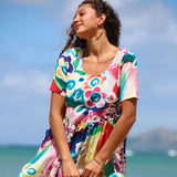 A woman wearing a colorful short-sleeve dress with a vibrant abstract floral pattern, standing outdoors against a blue sky and ocean background.