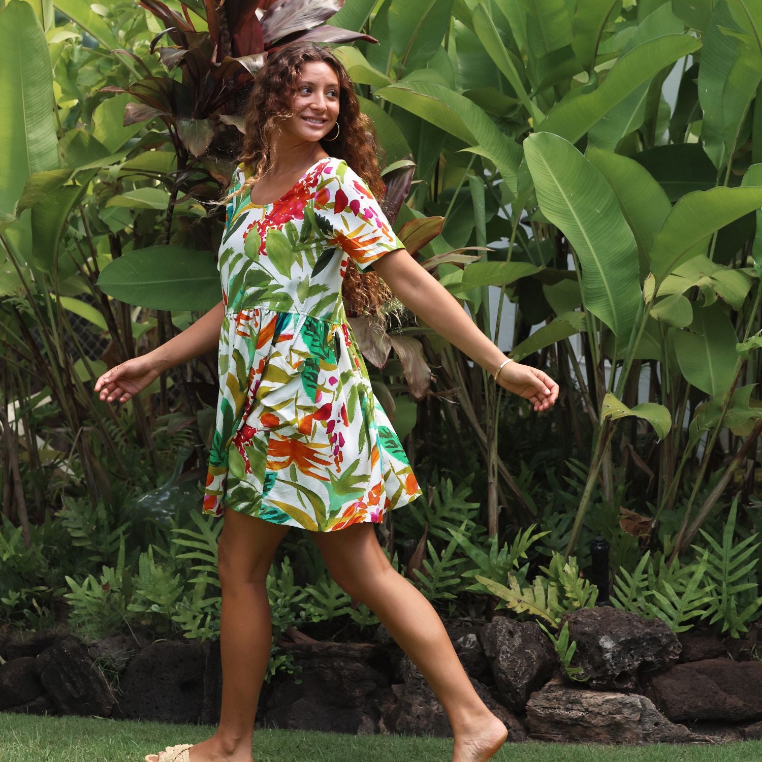 A woman wearing a short-sleeve white tropical floral print dress walks outdoors on a grassy lawn in front of a dense background of green tropical plants.