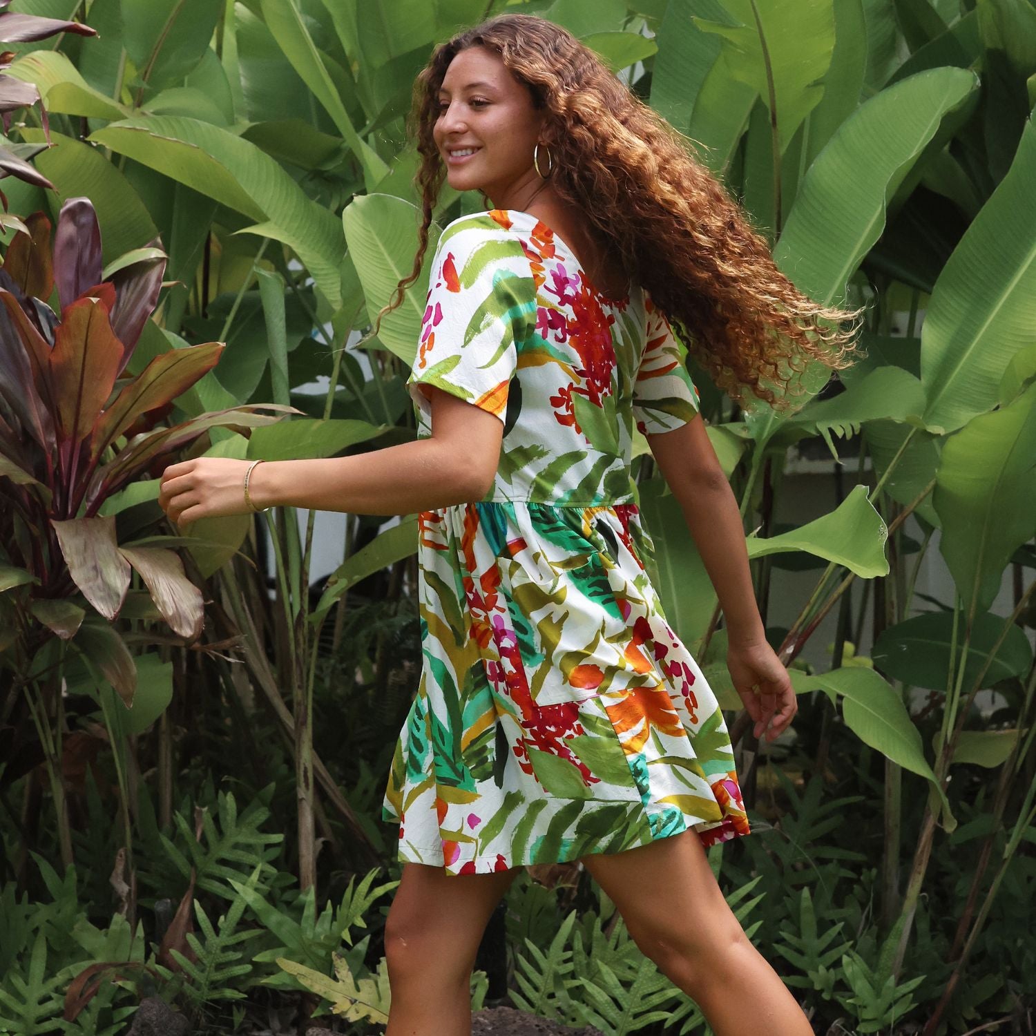 A woman wearing a short-sleeve white dress with a vibrant tropical leaf and floral print in green, red, orange, and yellow, walking through a lush tropical garden.