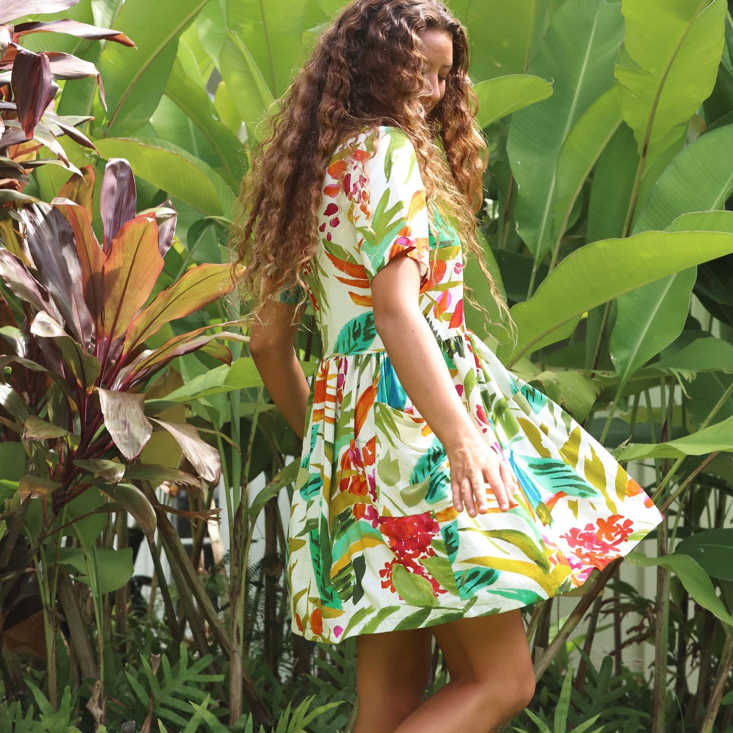 A woman wearing a short-sleeve white dress with a colorful tropical floral and leaf print, standing in a lush garden with large green leaves.