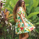 A woman wearing a short-sleeve white dress with a colorful tropical floral and leaf print, standing in a lush garden with large green leaves.