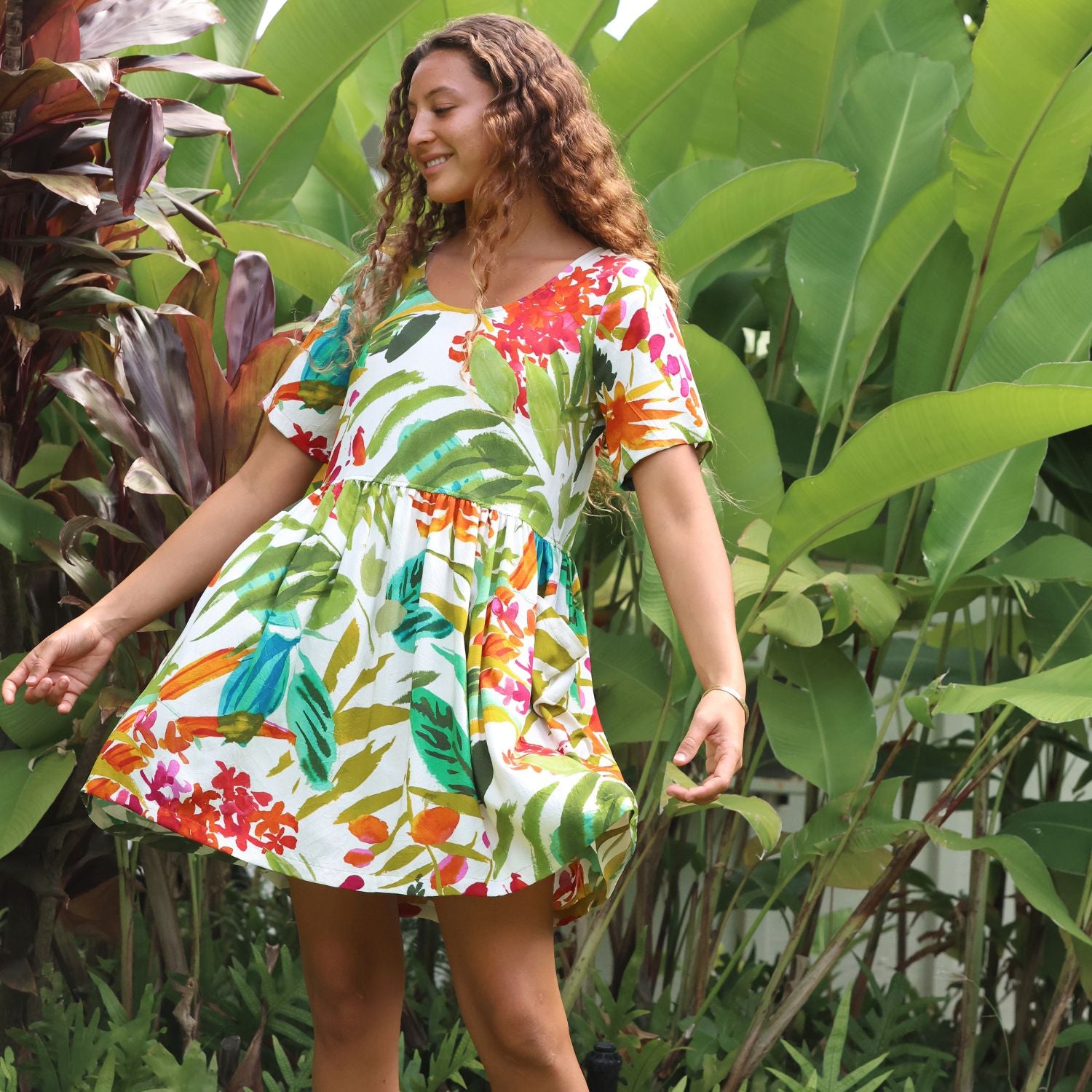 A woman wearing a short-sleeve white dress with a colorful tropical floral and leaf print, standing in a lush garden with large green leaves.