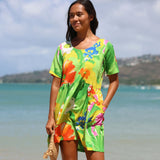 A woman wearing a vibrant tropical floral print mini dress stands on a beach with the ocean and distant hills in the background. The dress features a bright green, yellow, orange, and blue floral pattern.