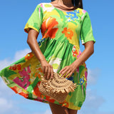 A woman wearing a bright green short-sleeve dress with a colorful floral print, holding a woven straw bag against a blue sky background.