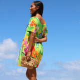 A woman wearing a bright multicolor floral off-the-shoulder dress and holding a woven straw bag, photographed outdoors against a clear blue sky.