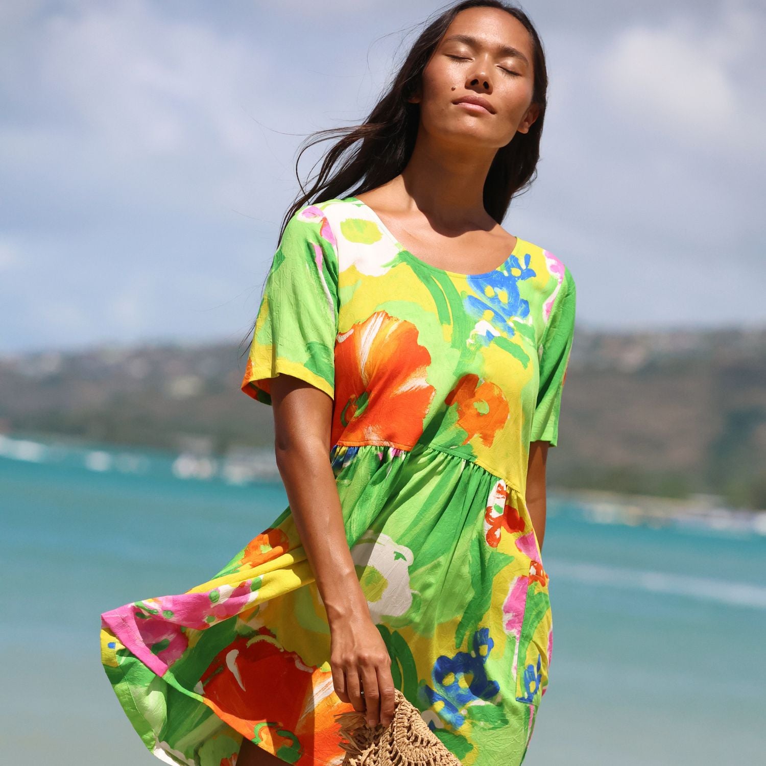 A woman wearing a bright tropical floral dress stands on a beach with the ocean and distant land visible in the background. The dress has a vivid green base with large orange, pink, yellow, blue, and white floral motifs.