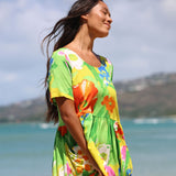 A woman wearing a bright tropical floral dress stands outdoors near the beach, with ocean water and hills blurred in the background.