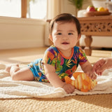 A baby wearing a colorful tropical print short-sleeve romper lies on a cream textured blanket on a woven rug, holding an orange and yellow striped fish toy. A hand is gently touching the toy.
