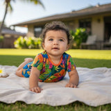 A baby lying on a white blanket on a grassy lawn, wearing a colorful tropical leaf print romper with a bright orange collar. The background shows a sunny backyard with a house and palm tree.