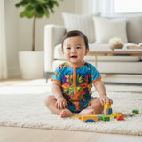 A baby sitting on a white rug in a bright living room, wearing a colorful tropical print one-piece swimsuit or romper. The swimsuit is bright blue with orange trim and a multicolor floral/tropical pattern.