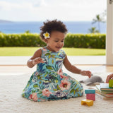 A toddler girl wearing a sleeveless turquoise floral dress sits on a white rug indoors, smiling and playing with colorful blocks and a gray plush toy. A real home interior and outdoor garden view are visible in the background.