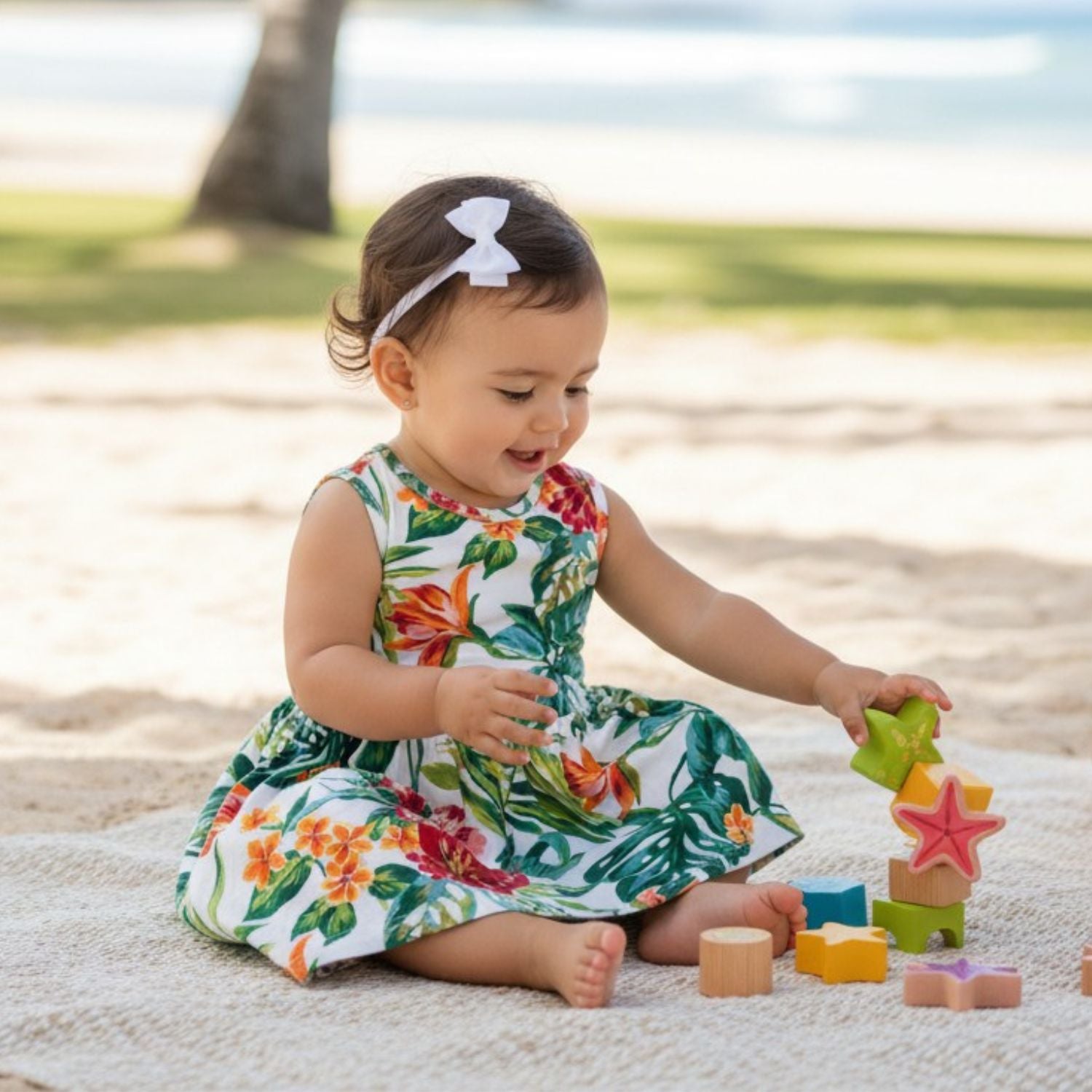 A toddler girl sitting on a blanket at the beach, wearing a white sleeveless dress with a colorful tropical floral print and a white bow headband, playing with wooden blocks.
