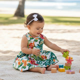 A toddler girl sitting on a blanket at the beach, wearing a white sleeveless dress with a colorful tropical floral print and a white bow headband, playing with wooden blocks.