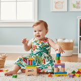 A baby girl sitting on a white carpet in a playroom, wearing a sleeveless tropical print dress with green palm leaves and red floral patterns. She is surrounded by colorful wooden building blocks.