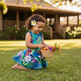 A young child kneeling on a grassy lawn in front of a wooden house, wearing a colorful tropical print sleeveless dress and holding a small woven basket with flowers.