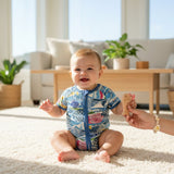A smiling baby sitting on a fluffy cream rug, wearing a blue short-sleeved romper with a colorful fish pattern.