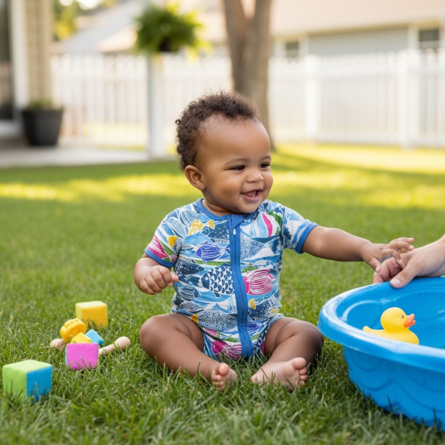 A smiling baby with short, curly dark hair sits on a green grass lawn wearing a blue, short-sleeved swimsuit covered in a colorful fish pattern. 