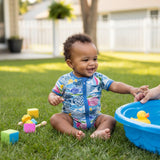 A smiling baby with short, curly dark hair sits on a green grass lawn wearing a blue, short-sleeved swimsuit covered in a colorful fish pattern. 