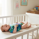A baby wearing a vibrant tropical-print onesie lies safely on its back inside a white wooden crib.