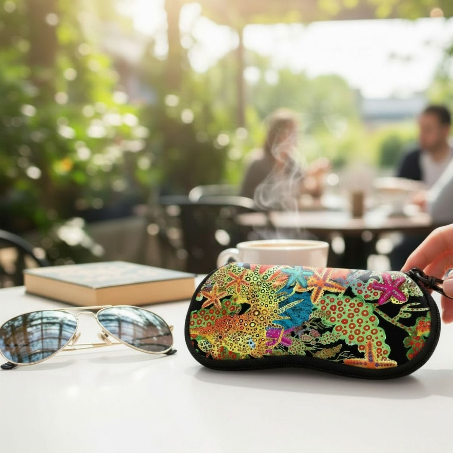 A black travel eye mask with a vibrant, multicolored coral reef and starfish pattern, held by a hand on a white table next to sunglasses and a book in an outdoor cafe setting.