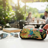 A black travel eye mask with a vibrant, multicolored coral reef and starfish pattern, held by a hand on a white table next to sunglasses and a book in an outdoor cafe setting.