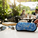 A close-up of a hand holding a blue and white tropical patterned zippered case on a white table, with aviator sunglasses and a blurred coffee cup in the background at an outdoor cafe.