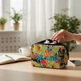 A colorful floral-patterned zippered pouch sits on a wooden desk with a hand placing white earbuds into it. The background shows a softly blurred home office setting with a cup, notebook, and plants.